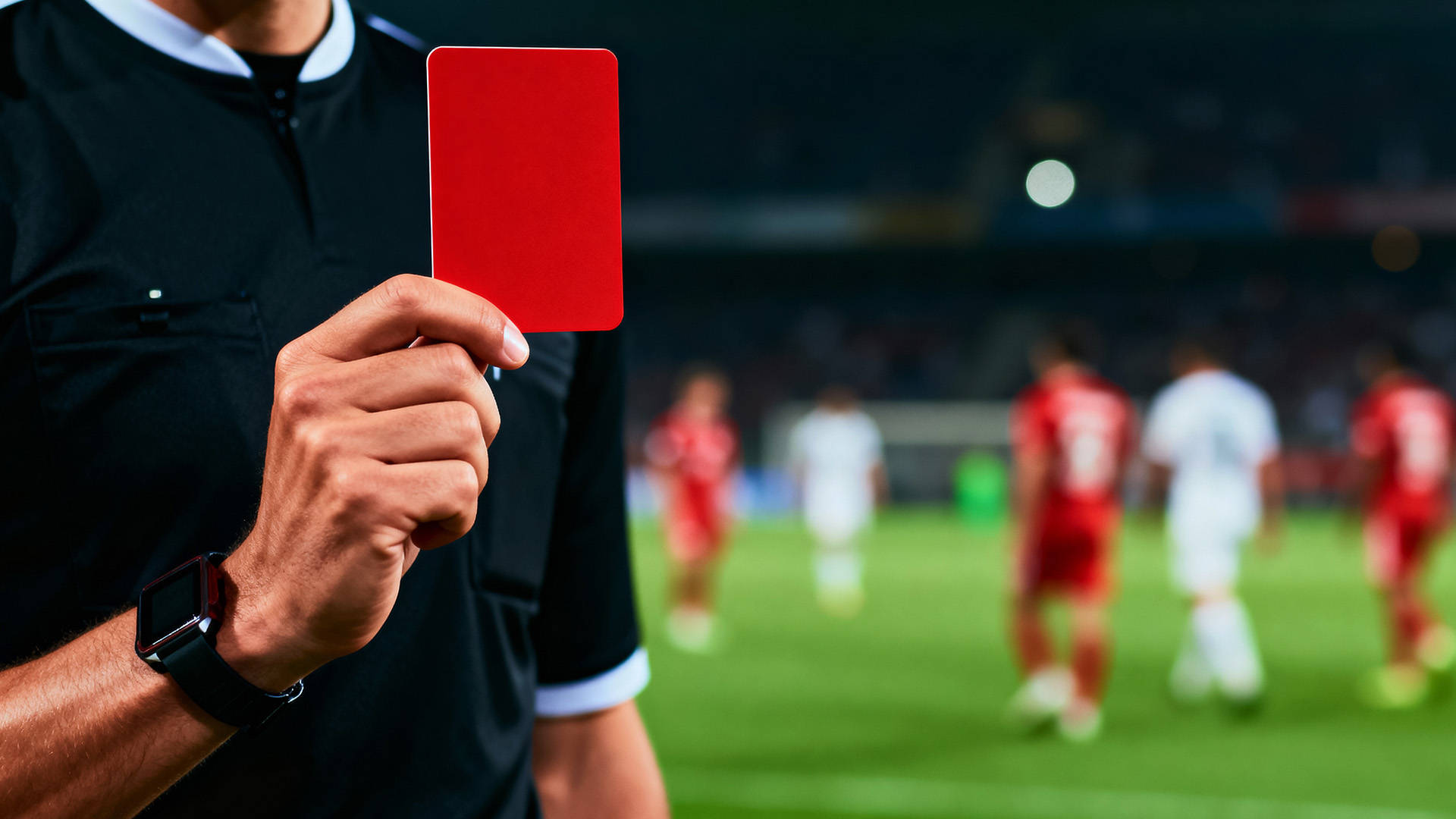 Dommer i sort uniform holder et rødt kort op på en fodboldbane, mens spillere ses sløret i baggrunden.
